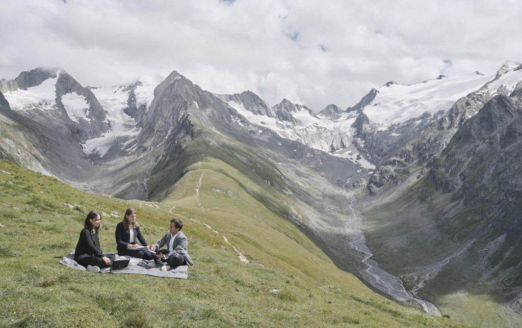 Drei Leute sitzen im Businessoutfit auf einer Picknickdecke in den Bergen
