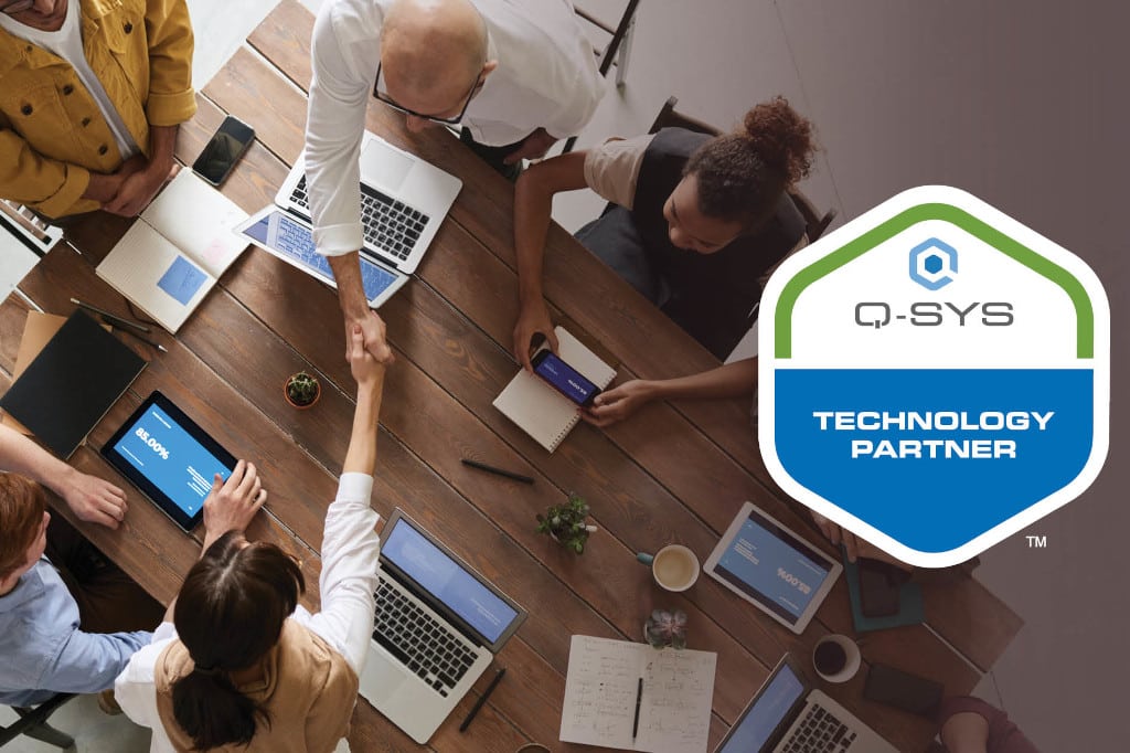 Aus der Vogelperspektive: Menschen schütteln sich die Hände und sitzen an Tisch mit Laptops; Q-SYS Technology-Partner-Program-Logo rechts im Bild