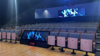 Blick auf Sitzreihen, mit einem kleinen Bildschirm in der ersten Reihe und einem sehr großen an der Wand