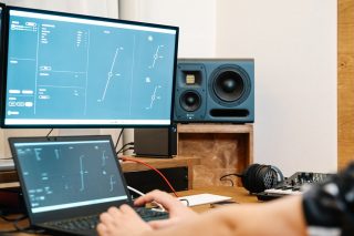 Studio-Szenerei mit zwei Monitoren, Lautsprechern und einem Mann davor, von dem nur die Hände zu sehen sind