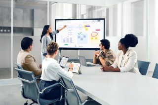 Besprechungsraum mit fünf Personen. Eine junge Frau zeigt auf ein Viewboard und dessen Inhalt