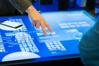 Arm eines Mannes mit zwei ausgestreckten Fingern über einem blauen Touchtisch schwebend