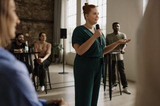 Frau mit Mikrofon steht in einem Raum, umgeben von Personen auf Barhockern