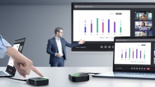 Mann an großem Bildschirm, vor ihm ein Besprechungstisch mit Notebook und Zubehör