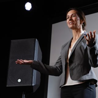 Frau mit ausgebreitetem Händen, neben ihr ein schwarzer Lautsprecher