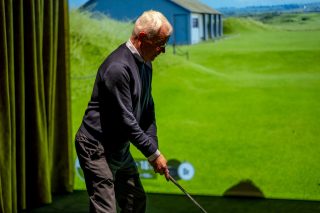 Älterer Mann schwingt Golfschläger vor Wand mit Projketion eines Fairways