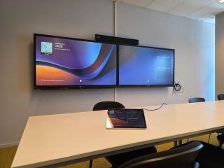 Zwei Monitore nebeneinander an einer Wand vor einem langen Tisch, auf dem ein Tablet liegt
