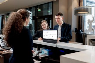 Pärchen an Service-Desk und Mitarbeiterin schauen auf den beidseitigen Bildschirm