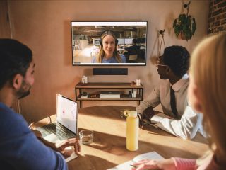 Zwei Menschen in kleinem Meeting-Raum an einem Tisch und eine Display mit Gesprächspartnerin an der Wan