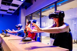 Drei Personen mit Vr-Brillen an einem Tisch nebeneinander, die Hände ausgestreckt