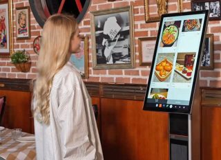 Frau mit bloneden langen Haaren vor großem hochkantigen Touchsscreen mit Bildern von Essen