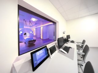 Bedienplatz mit mehreren Monitoren und einem Touchpanel, durch ein großes Sichtfenster ein violett beleuchteter MRT-Raum mit Untersuchungsgerät.