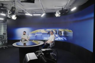 Zwei Männer sitzen lässig an einem rund Tisch vor einer gebogenen LED-Wand in einem ganz in blau gehaltenen Ambiente