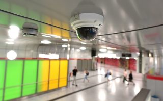 Weiße Dome-Überwachungskamera an der Decke einer Stadtbahn-Station, im unscharfen Hintergrund eine Verteilerebene mit grün-gelber Wandgestaltung und mehreren Passanten.