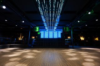 Leere Tanzfläche mit Holzboden in einem Club, an der dunklen Decke geflogene Lautsprecher und Scheinwerfer, im Hintergrund ein blau beleuchteter DJ-Bereich