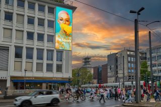 Auffälliger Großbildschirm an Hausfassade zeigt Frauenkopf mit Brille und gelbem Schaum