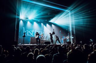 Vierköpfige Band auf einer Bühne im blauen Scheinwerferlicht, im Hintergrund eine Projektion mit Bandnamen, davor dichtes Publikum