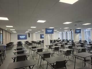 Großer Lehrsaal mit Reihen leerer Einzeltische und Stühlen, an Wänden und Säule mehrere Bildschirme mit Uhrzeitanzeige, darüber Deckenlautsprecher und Rasterleuchten.