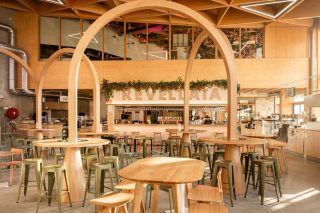 Gastraum mit hohen Holzbögen und Stehtischen, im Hintergrund eine Bar mit dem beleuchteten Schriftzug Revellata und Begrünung über der Theke.