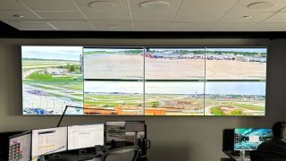 Kontrollraum mit einer Videowand aus acht Displays, die Vorfeld, Rollwege und Terminalgebäude eines Flughafens zeigen, davor Arbeitsplätze mit mehreren Monitoren.