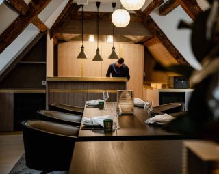 Gedeckter Restauranttisch unter historischem Dachgebälk, im Hintergrund ein Mitarbeiter an einer Holztheke mit Pendelleuchten