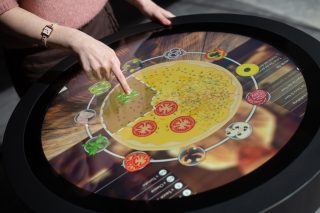 Runder Touchtisch mit schwarzem Rahmen, auf dem Display eine Pizza-Konfigurations-App mit kreisförmig angeordneten Zutatensymbolen, eine Hand tippt auf ein Symbol.