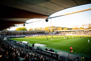 Blick unter dem Tribünendach hindurch auf ein laufendes Fußballspiel im BUKO Stadion bei tief stehender Sonne, unter dem Dach sind Lautsprecher montiert, im Vordergrund Zuschauer und ein Linienrichter