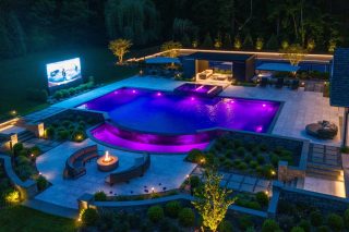 Nächtliche Luftaufnahme eines Anwesens mit violett beleuchtetem Pool, Feuerstelle, überdachter Sitzgruppe und einer großen Videowand am Rand der Terrasse.