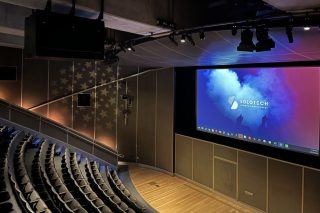 _: Blick von den oberen Sitzreihen des Leonard Nimoy Event Horizon Theater auf Bühne und Projektionsleinwand, an der Decke Scheinwerfer und ein schwarzes Gerätegehäuse, an der Seitenwand ein Sternenmuster
