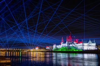 Parlamentsgebäude in Budapest bei Nacht in Rot, Weiß und Grün angestrahlt, darüber blaue Laserstrahlen
