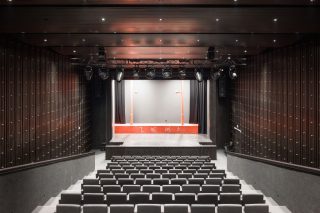 Großer Saal des Divadlo Poprad mit ansteigenden grauen Sitzreihen, dunklen Wänden, Lichttraverse und roter Bühnenkante