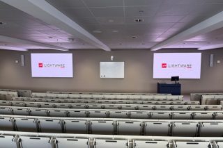 Blick von den hinteren Sitzreihen auf einen Hörsaal mit zwei großen Displays, Whiteboard und Rednerpult.