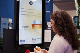 Eine Frau hält ein Kosmetikprodukt vor einem digitalen Display, auf dem Informationen zum Produkt angezeigt werden.