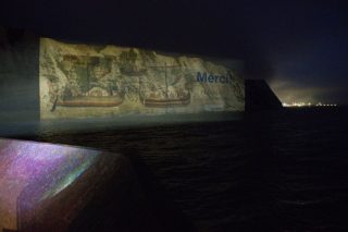 Nächtliche Projektion auf den Kreidefelsen von Dover mit historischen Bildmotiven, dem Schriftzug „Merci!“ und dem Logo des British Museum