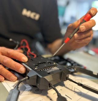 Zwei Hände öffnen mit einem Schraubendreher ein schwarzes Kameramodul auf einer Werkbank, im Hintergrund ein Mitarbeiter im MCI-Shirt