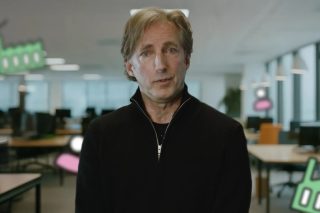 Stephen Fuller in schwarzem Pullover mit Reißverschluss in einem Großraumbüro, umgeben von grafischen Symbolen für Fabriken und Personen