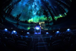 Innenraum des Zeiss-Planetariums Jena mit voll besetzter Projektionskuppel. Darauf ist ein Sternenhimmel mit grünem Polarlicht über einer Waldsilhouette zu sehen. In der Saalmitte steht der beleuchtete Sternenprojektor, davor Sitzreihen