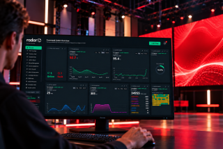 Person am Schreibtisch vor einem Monitor mit dem Radar-Dashboard, im Hintergrund eine Halle mit roter LED-Wand und Traversen