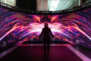 Eine Frau steht mit ausgebreiteten Armen vor einer gebogenen LED-Wand. Darauf ist eine leuchtende Grafik aus pinken, orangen und blauen Lichtlinien zu sehen, die an Flügel erinnert. Darüber verläuft eine Traverse mit Lautsprechern und Kameras.