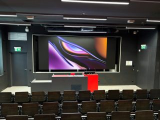 Hörsaal mit dunklen Wänden, Stuhlreihen und Rednerpult, an der Stirnwand eine breite LED-Wand mit abstrakter Grafik in Violett und Orange