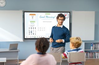 Ein Lehrer mit Brille und blauem Pullover steht in einem Klassenzimmer neben einem großen Touchdisplay zwischen zwei aufgeklappten Whiteboard-Flügeln. Auf dem Display ist eine Unterrichtsoberfläche zu sehen, mit Kalenderblatt, der Begrüßung „Good Morning!“, Timer und Formenauswahl. Im Vordergrund sitzen zwei Kinder, von hinten fotografiert