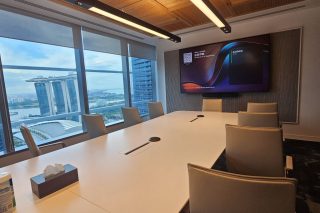 Konferenzraum mit langem weißem Tisch und hellen Lederstühlen. An der Stirnwand hängt ein großes Display mit dem Microsoft-Teams-Rooms-Startbildschirm, darunter eine Videokonferenz-Bar. Durch die Fensterfront ist eine Hochhaus-Skyline am Wasser zu sehen.