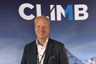 Mann im dunklen Sakko mit Climb-Umhängeband vor einer Fotowand mit Climb-Logo und schneebedeckten Bergen