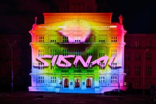 Klassizistische Gebäudefassade bei Nacht, mit bunten Farbverläufen und dem Schriftzug „Signal“ angestrahlt