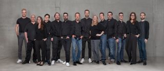 Gruppenfoto von 14 Personen in überwiegend schwarzer Kleidung vor einer grauen Betonwand