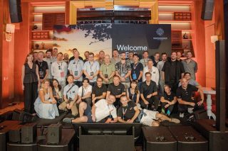 Gruppenfoto mit rund 40 Personen mit bunten Konferenz-Lanyards auf und vor einer Bühne. Dahinter eine LED-Wand mit Sonnenuntergangsmotiv, Begrüßungsschriftzug
