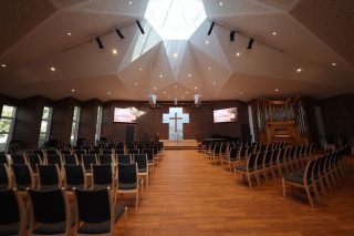 Kirchensaal mit Stuhlreihen, Holzboden und einer zeltartigen Decke mit Oberlicht, vorne ein beleuchtetes Kreuz, zwei Displays, eine Orgel und zwei abgehängte weiße Lautsprecher