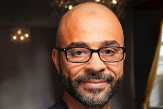 Porträt von Mo Gawdat mit Glatze, Brille und grauem Bart vor dunklem Hintergrund mit Kronleuchter