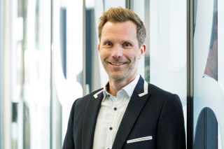 Portrait von Messedirektor Maik Fischer. Lächelnder Mann mit kurzen blonden Haaren, weißem Hemd und dunklem Sakko vor einer Glaswand.
